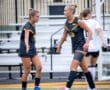 Two female soccer players in black uniforms discussing a play on the field, with empty bleachers in the background.