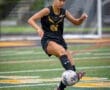 Soccer player in black jersey kicks a ball on a field during a game, showcasing athleticism and focus.