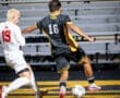 Soccer players compete on the field; one player kicks the ball while another attempts to defend.
