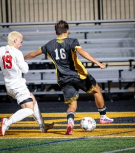Soccer players compete on the field; one player kicks the ball while another attempts to defend.