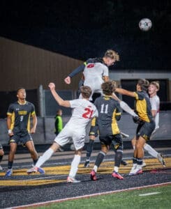 High school soccer players leap for the ball in a competitive match under the stadium lights, showcasing athleticism.