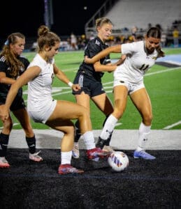 High school girls' soccer match action, featuring intense play as players compete for the ball on the field at night.