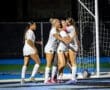 Four female soccer players in white jerseys celebrate a goal with joyful hugs on a night game field.