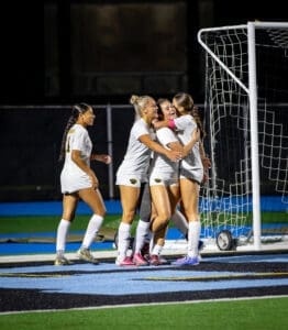 Four female soccer players in white jerseys celebrate a goal with joyful hugs on a night game field.