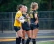 Three soccer players discuss strategy on the field, with the goalkeeper in yellow and two teammates in black uniforms.