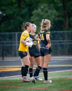 Three soccer players discuss strategy on the field, with the goalkeeper in yellow and two teammates in black uniforms.