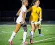 Two female soccer players celebrate on the field at night, showcasing teamwork and athletic spirit in their uniforms.