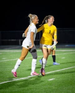 Two female soccer players celebrate on the field at night, showcasing teamwork and athletic spirit in their uniforms.