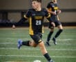 Soccer player in black and gold jersey dribbling a ball on a field during a game under stadium lights.