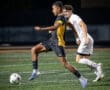 A player in a black and yellow soccer uniform dribbles the ball while being closely guarded by an opponent.