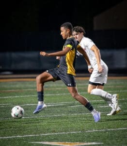 A player in a black and yellow soccer uniform dribbles the ball while being closely guarded by an opponent.