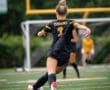 Female soccer player in black uniform kicking a ball on a field with goalposts in the background. Action shot.