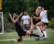 Two female soccer players compete for the ball, one sliding to tackle while others watch intently on the sidelines.