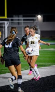 Two female soccer players compete for control of the ball during a night game on a well-lit field.