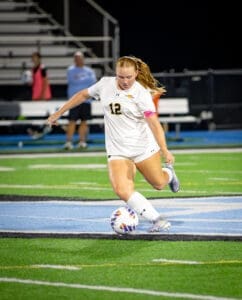 A soccer player in a white jersey kicks a ball on a field during a night game. Focused and determined.