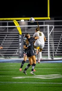 Two female soccer players compete for a high ball during a night game on a well-lit field.