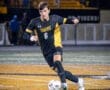 A soccer player in a black and yellow jersey dribbles a ball on the field during a night game.