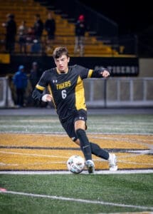 A soccer player in a black and yellow jersey dribbles a ball on the field during a night game.