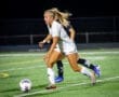 Female soccer player in white uniform sprinting after a ball during a night match on a well-lit field.