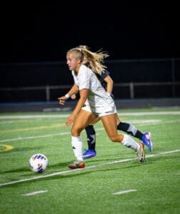 Female soccer player in white uniform sprinting after a ball during a night match on a well-lit field.