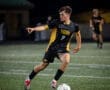 Soccer player in action under stadium lights, wearing a black and yellow jersey, dribbling a ball on the field.