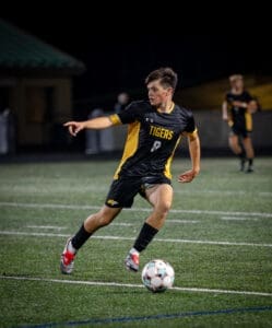 Soccer player in action under stadium lights, wearing a black and yellow jersey, dribbling a ball on the field.