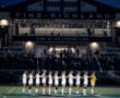 Pine-Richland soccer team standing in line during national anthem at night, with fans in the background at the stadium.