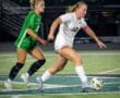 A soccer player in a white uniform dribbles the ball past a defender in a green jersey on a field at night.