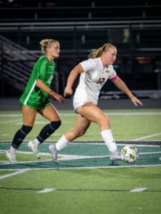 A soccer player in a white uniform dribbles the ball past a defender in a green jersey on a field at night.