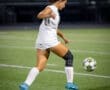 Female soccer player in action, dribbling the ball on a field at night with stadium lights illuminating the scene.