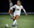 A soccer player in a white uniform takes a shot during a night game, showcasing strong athleticism on the field.