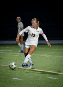 A soccer player in a white uniform takes a shot during a night game, showcasing strong athleticism on the field.