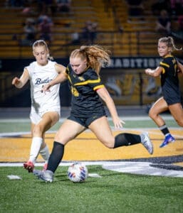 Two female soccer players competing for the ball on the field during a night game, with spectators in the background.