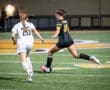 Two female soccer players sprinting on a field at night, one in a white uniform and the other in black.