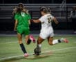 High school soccer match featuring players in green and white jerseys competing for the ball under stadium lights.