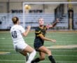 Two female soccer players competing on the field; one headlining the ball while the other defends. Night game action.