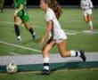 High school soccer match at night, featuring a player in white running for the ball, with teammates in the background.