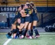 A group of female soccer players celebrates a goal, embracing each other joyfully on the field at night.