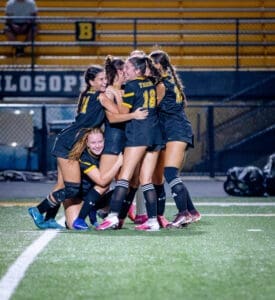 Girls' soccer team celebrating a goal, embracing in joy on the field under stadium lights. Team spirit and camaraderie evident.
