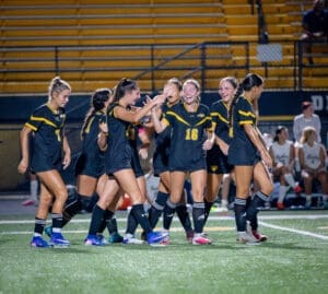 Girls' soccer team celebrating a goal under the night lights on the field, showcasing teamwork and excitement.
