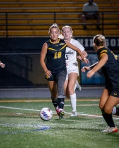 Female soccer players in action during a match, one dribbling the ball while others run behind on a lit field.