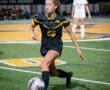 Female soccer player dribbling the ball on a field during a night game, wearing a black jersey with number 25.