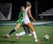 A player dribbles the soccer ball while a goalie in green prepares to defend during a night game on the field.