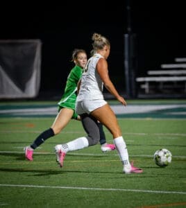A player dribbles the soccer ball while a goalie in green prepares to defend during a night game on the field.