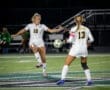 Two female soccer players in white uniforms engage in a game, one kicking a soccer ball on a field at night.