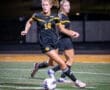 High school girls’ soccer players in black uniforms play under stadium lights, focused on the soccer ball on the field.