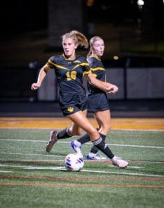 High school girls’ soccer players in black uniforms play under stadium lights, focused on the soccer ball on the field.
