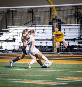 High school soccer match with players competing for the ball; a goalkeeper watches from the goal.