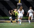 Soccer players in a tense moment, with the goalkeeper diving to block a shot at night on a well-lit field.