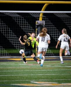 Soccer players in a tense moment, with the goalkeeper diving to block a shot at night on a well-lit field.